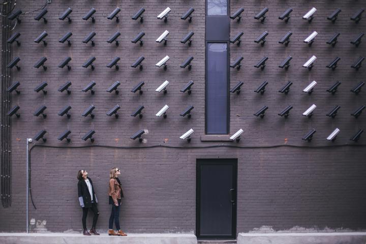 Outside wall with many security cameras pointing down at 2 people at a door